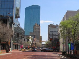 A city street with red brick pavement, lined with trees and modern buildings. A tall, glass skyscraper and several older buildings stand in the background under a clear blue sky. Cars are stopped at a traffic light.