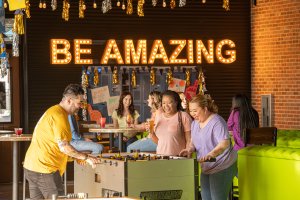 People gather in a lively room, playing foosball around a table in the foreground. In the background, others chat at a high table. A bright sign with lit-up letters spells out "BE AMAZING." The atmosphere is vibrant and cheerful, with festive decorations hanging above.