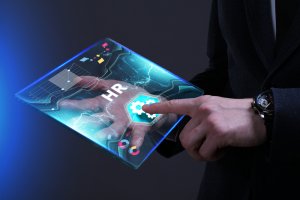 A person in a dark suit is holding and interacting with a futuristic, transparent tablet displaying HR trends and related icons. The screen shows graphs, a gear icon labeled "HR," and various digital elements in a blue, glowing interface.