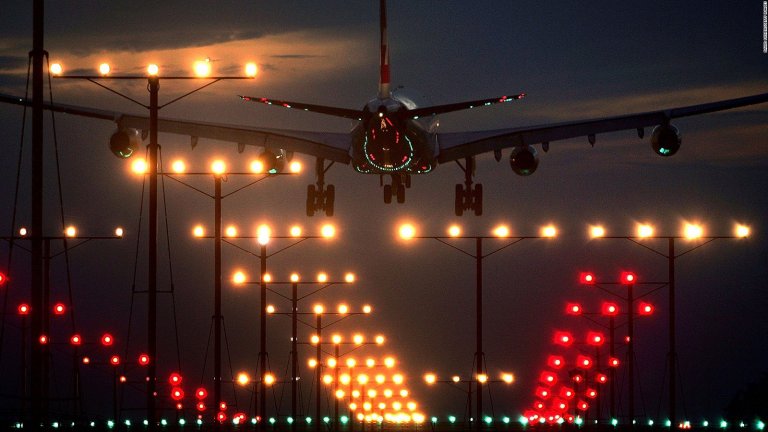 A large airplane approaches a runway at night at LAX, surrounded by numerous bright airport landing lights glowing in white, red, and green. The sky is dark with a hint of twilight, creating a dramatic contrast with the illuminated path guiding the aircraft's descent—a striking LAX aviation scene.