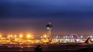 A brightly lit LAX aviation terminal at night, showcasing several aircraft parked on the tarmac. The control tower is prominent in the background, with the sky transitioning from dark to lighter hues. The lights from the terminal and planes create a vibrant glow.
