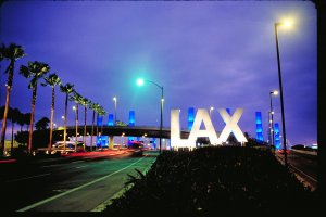 A night view of the LAX airport sign, illuminated in white with blue light columns in the background. Surrounding the sign are tall palm trees, streetlights, and a busy street with blurred car lights, indicating moving traffic. The sky is a deep purple—a testament to Los Angeles contractors' stunning cityscape designs.