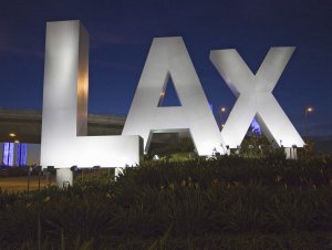 Large illuminated letters "LAX" stand prominently in front of lush greenery at night, symbolizing the heart of LAX aviation. The sky is dark, with dimly lit streetlights and a bridge in the background.