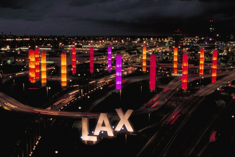 A nighttime view of the entrance to Los Angeles International Airport (LAX) showcases tall, illuminated pylons in vibrant colors like orange, pink, and purple. The pylons line the roads leading to the airport, with the iconic "LAX" letters prominently displayed in the foreground.