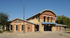 A modern, two-story public library building with red brick walls, an arched roof section, and a blue awning over the entrance. The sign reads "John and Judy Gay Library," and there is a parking lot in the foreground with a lamppost on the left. Trees surround the area.