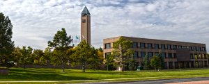 A large clock tower stands tall beside a multi-story brick building with a triangular roof. The building is surrounded by trees and a well-manicured lawn under a partly cloudy sky. Flags are seen near the tower, indicating it may be an important or official site.