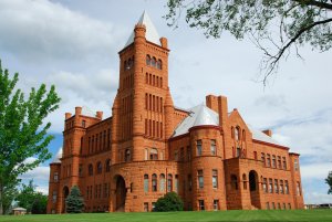A large, historic red sandstone building with a tall central tower, arched windows, and a steeply pitched roof. The structure is surrounded by well-maintained green lawns, with trees on either side and a partly cloudy sky above.