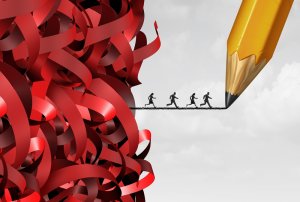An illustration shows four silhouetted individuals running on a platform drawn by a giant pencil. They are heading towards a tangled mass of red tape on the left side, symbolizing bureaucracy and obstacles. The background transitions from grey to white.