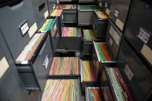 An organized filing system with multiple grey drawers, each containing rows of color-coded folders. The drawers are open, revealing neatly arranged folders in shades of green, yellow, pink, and white. Labels are affixed to the front of the drawers, maintaining a semblance of order amidst bureaucracy.