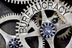 Close-up image of interlocking metal gears with the word "BUREAUCRACY" engraved on one. The intricately connected gears symbolize the complex and intertwined nature of bureaucracy.