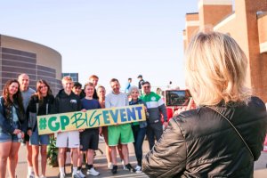 A group of people pose outdoors, smiling and holding a large "#GPBIGEVENT" sign, while someone in a black jacket takes their photo with a smartphone. Modern buildings are visible in the background.