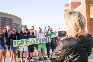 A group of smiling people poses outdoors holding a large sign that says "#GPBIGEVENT." A person in the foreground with blonde hair and a black jacket takes a photo of the group using a smartphone. The event appears to be taking place near large buildings.