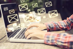 A person wearing a plaid shirt types on a laptop, while floating envelope icons symbolize incoming emails. The casual indoor setting features a wooden table, creating a cozy and focused workspace.