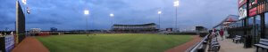 Panoramic view of a baseball stadium during sunset, with a few players on the field. The stadium is partially filled with spectators. The scoreboard and seating area are visible on the right, and the American and Texas flags are flying on the left.