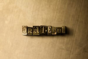 The word "GRATEFUL" is formed using old metal letterpress type blocks, arranged in a horizontal line on a textured, sepia-toned background. Each letter block features a different style, adding a vintage feel to the composition and evoking a deep sense of gratitude.