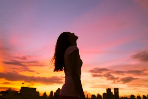 A silhouette of a woman with long hair stands outdoors at sunset, looking up towards the sky in gratitude. The skyline of a city with buildings can be seen in the background, against a colorful sky painted in shades of orange, pink, and purple.