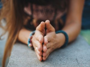 Close-up of a person's folded hands resting on a concrete surface, exuding a sense of gratitude. The person is wearing bracelets on both wrists and has long hair. The background is blurred, focusing on the hands in the praying position.