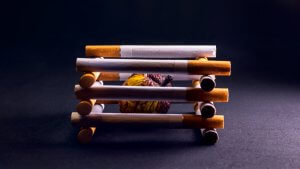 A dark background with a close-up of several cigarettes arranged in a cage-like structure. Inside is a shriveled, darkened piece of organic matter, encapsulating the destructive habit of smoking. The image delivers a powerful visual metaphor for its harmful effects.