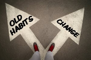 A person stands at a fork in the road with two large arrows painted on the ground. The left arrow labeled "Old Habits" points left, and the right arrow labeled "Change" points right. The habit-clad person's feet, in red-laced shoes, are visible at the junction.