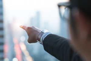 Close-up of a leader wearing glasses and a business suit, pointing towards a blurred urban cityscape with high-rise buildings. The background appears foggy or out-of-focus, and the individual's watch is visible on their wrist.