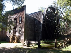 A large, old wooden watermill with a tall water wheel stands next to a split-rail fence, surrounded by trees and sunlight. Bundles of corn stalks and pumpkins decorate the entrance.