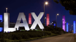 A nighttime view of the iconic large "LAX" letters at the entrance of Los Angeles International Airport. The letters are brightly lit, and tall towers in the background are illuminated with colorful red, blue, and purple lights. The street beside LAX is empty.
