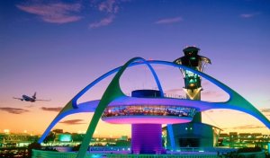 A futuristic structure with neon lights at dusk, featuring a tall central column and sweeping arches resembling a spider or spaceship, reminiscent of LAX. An airplane is seen in the sky to the left and an illuminated cityscape lies in the background.