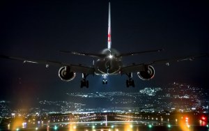 A commercial airplane descends towards a runway at LAX at night, with its landing gear extended and lights illuminating the path. The cityscape in the background is dotted with lights, accentuating the dark sky. The plane's engines and wings are clearly visible.