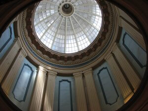 A circular domed ceiling with an intricate pattern of glass panes allowing natural light to flood in. The dome is supported by tall, elegant columns and features ornate architectural details. The light casts soft shadows, highlighting the grandeur of the structure.