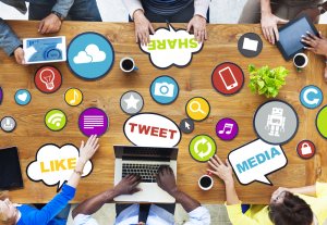 A group of people sit around a wooden table, engaging with various social media icons such as "Tweet," "Like," and "Share." The table is covered with diverse symbols representing social media platforms and digital communication tools. Each person uses a device.