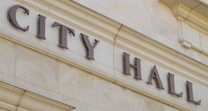 Close-up view of a beige stone building with the words "City Hall" prominently displayed in large, dark letters. The architectural details include a ledge and decorative molding.
