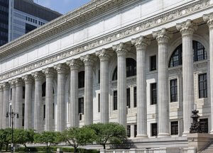 A large neoclassical building features a series of tall Corinthian columns supporting a detailed frieze with classical motifs. Arched windows are evenly spaced along the facade, and manicured landscaping with trees and bushes can be seen in the foreground.