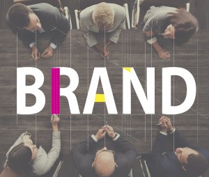A group of six people, dressed in business attire, sit around a wooden table with the word "BRAND" superimposed over them in large, bold letters. The letters are stylized with design guidelines and color accents. The image suggests a branding meeting or discussion.