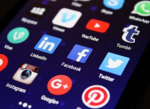 A close-up of a smartphone screen showing various social media app icons, including LinkedIn, YouTube, Twitter, Facebook, Instagram, Tumblr, Pinterest, Google+, and Vimeo against a dark background.
