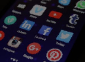 A blurry close-up image of a smartphone screen displaying various social media app icons, including Instagram, Google+, Pinterest, LinkedIn, Facebook, YouTube, Twitter, and others. The background is dark, and the icons are in different colors.