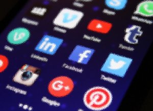 Close-up image of a smartphone screen displaying various social media app icons, including LinkedIn, Facebook, Twitter, Instagram, Google+, Pinterest, Tumblr, and Vine. The background is slightly blurred, creating a textured and artistic effect.