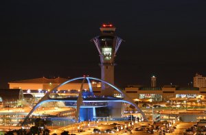 A nighttime view of an illuminated airport featuring a distinctive futuristic structure with arching white beams in the foreground and a control tower in the background, expertly crafted by Los Angeles contractors. The surrounding area is filled with lights from nearby buildings and parked cars.
