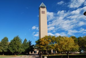 A tall brick clock tower with a pointed metal roof stands against a bright blue sky dotted with clouds. The tower is surrounded by green and yellow-leaved trees, and a well-maintained lawn is visible in the foreground.