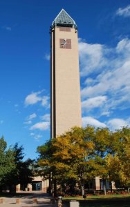 A tall clock tower with a triangular glass top stands against a bright blue sky with scattered clouds; trees with green and yellow leaves are in the foreground.