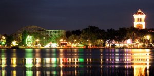 Nighttime view of an amusement park with vibrant, colorful lights reflecting on a calm body of water. A roller coaster and a brightly lit tower are visible in the background, surrounded by trees. The scene exudes a lively and festive atmosphere.