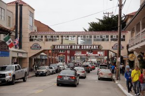 A busy street scene on Cannery Row in Monterey, California, with cars, pedestrians, and a bridge overhead displaying the "Cannery Row Company" sign. Buildings and flags line the street.