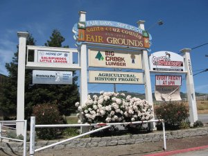 Large outdoor sign for Santa Cruz County Fairgrounds, advertising Fair Days (Sept 15-20), Big Creek Lumber, Ocean Speedway, Little League, and the Agricultural History Project. White flowers grow at the base.