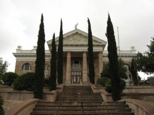 A stone courthouse with tall columns and decorative pediment, partially obscured by tall, narrow cypress trees lining a wide staircase leading to the entrance.