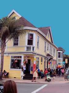 A yellow, two-story building with shops and people gathered outside. Some hold balloons; a woman pushes a stroller while others chat or sit on benches. Palm tree on the left, blue sky in the background.