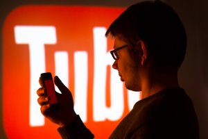 A person wearing glasses looks at their smartphone while standing against a large, illuminated YouTube logo in the background. The image, steeped in a business-like ambiance, is dark except for the light from the logo and phone screen.