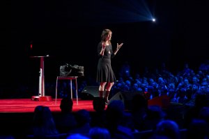 A woman stands on a red-lit stage delivering a speech to a seated audience at a formal business event. She gestures with her hands, wearing a black dress and boots. A podium and black bag are placed beside her on the stage as a spotlight shines on her from above.