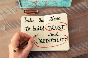 A hand holding a pen writes in an open notebook. The words "Take the time to build TRUST and CREDIBILITY" are written in black ink, with "TRUST" and "CREDIBILITY" circled in red. The notebook rests on a wooden surface with a turquoise decorative item visible in the background, emphasizing credibility.