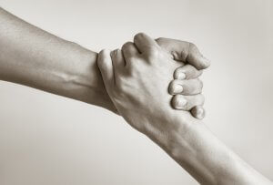 Two hands are clasped tightly in a firm grip, symbolizing support and generosity. The image features a close-up view with one hand reaching up while the other reaches down, creating a sense of connection and mutual assistance. The background is neutral.
