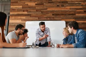 A group of five people is seated around a table in a meeting room, attentively listening to a man standing and speaking with generosity. Behind him is a whiteboard on a wooden wall. The table holds notebooks, pens, a pair of glasses, and a glass of water.