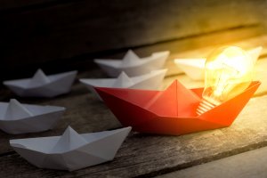 A red origami boat with an illuminated light bulb inside it is surrounded by several white origami boats on a wooden surface. The light bulb emits a warm glow, highlighting the red boat's generosity among the white ones.